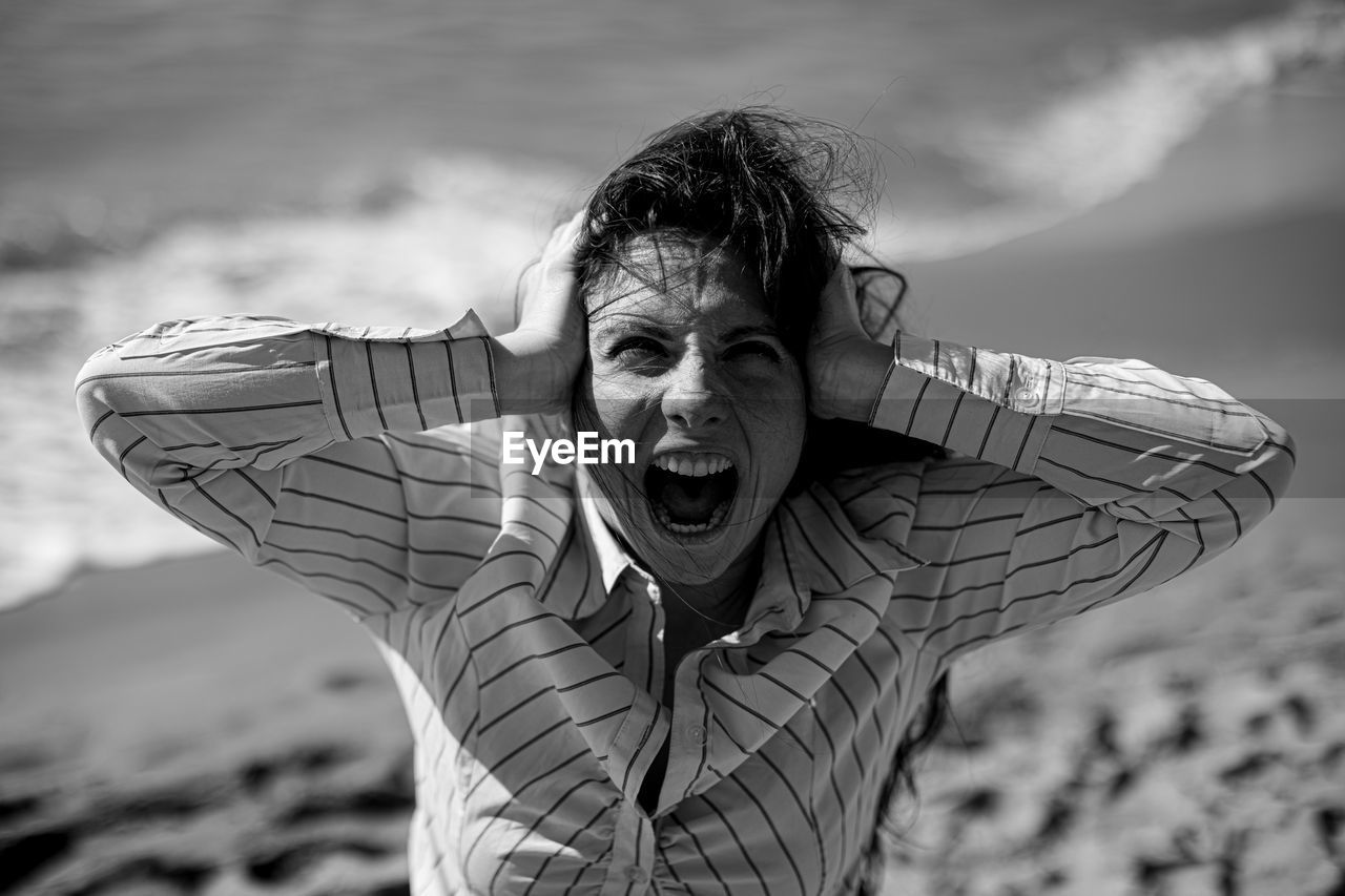 Portrait of frustrated woman with hands covering ears shouting at beach against sky