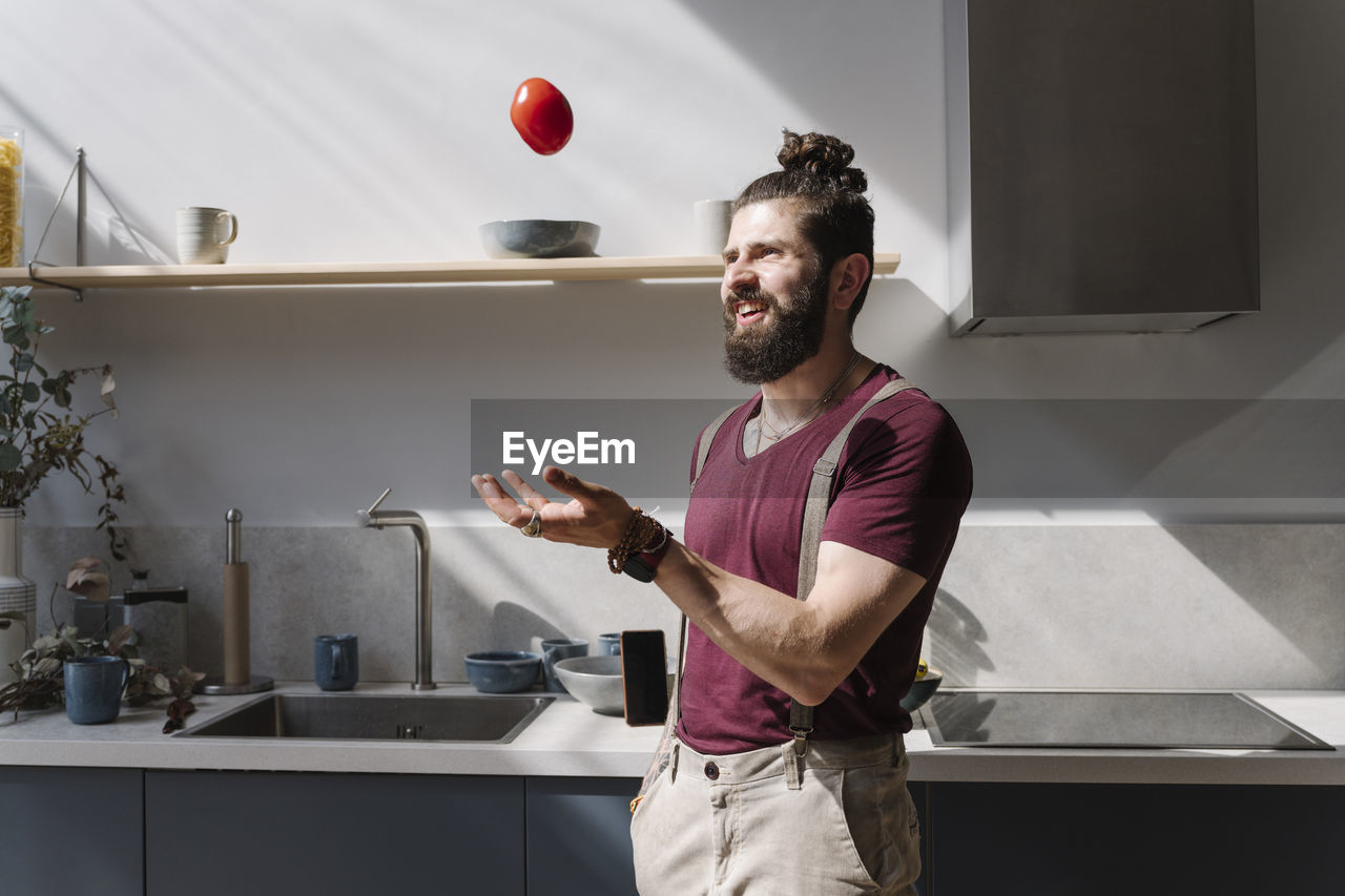 Bearded man playing with tomato while standing at kitchen counter