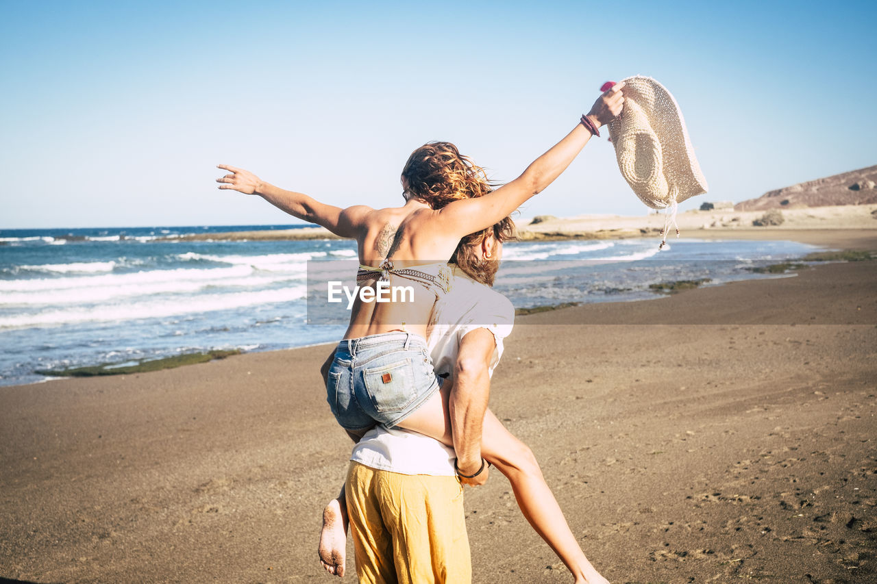 Rear view of man piggybacking woman at beach against clear sky