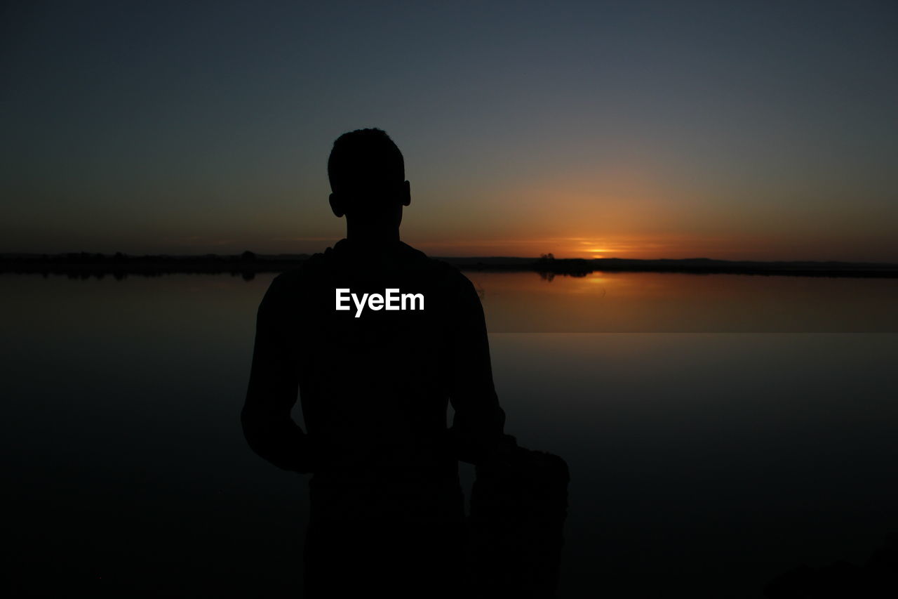 Silhouette man standing by lake against sky during sunset