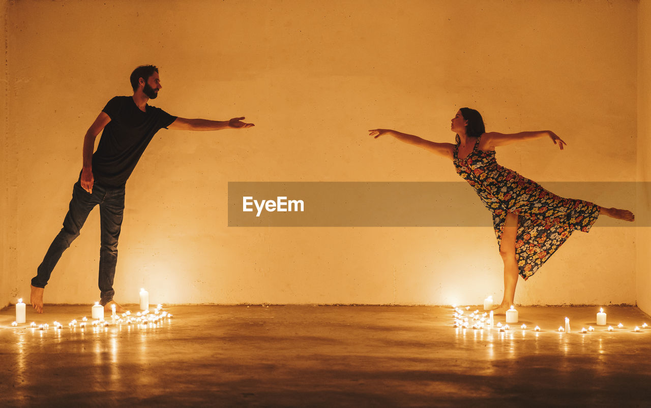 Couple stretching arms to reach while standing amidst lit candles in darkroom