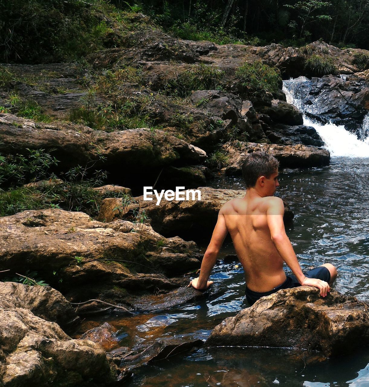 Rear view of shirtless man sitting on rock by stream