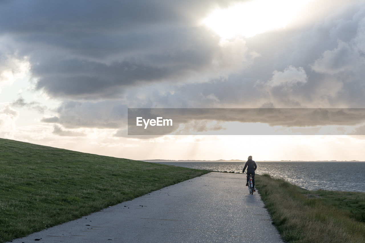 Rear view of man riding bicycle on road by sea against cloudy sky