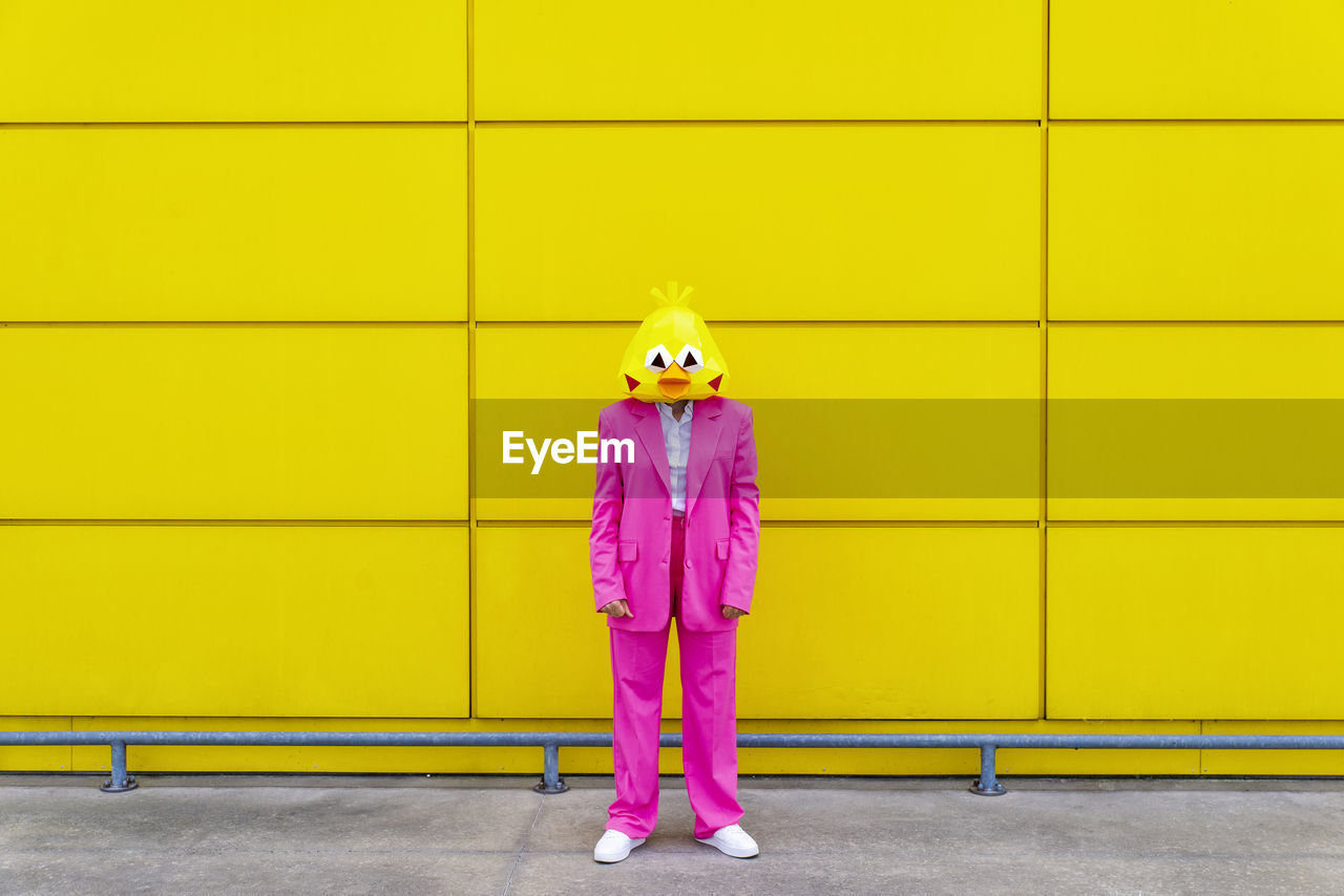 Woman wearing vibrant pink suit and bird mask standing in front of yellow wall