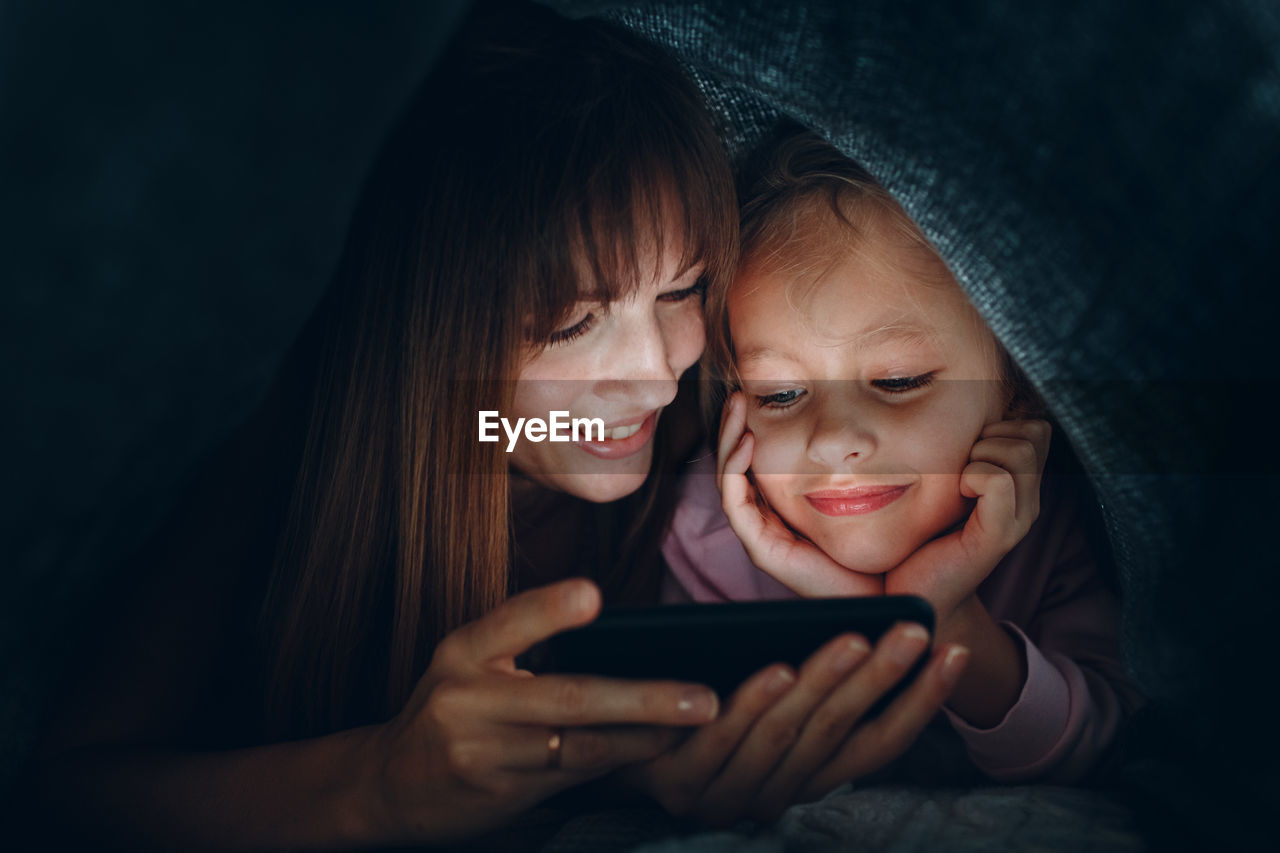 Smiling mother and daughter looking at mobile phone under blanket