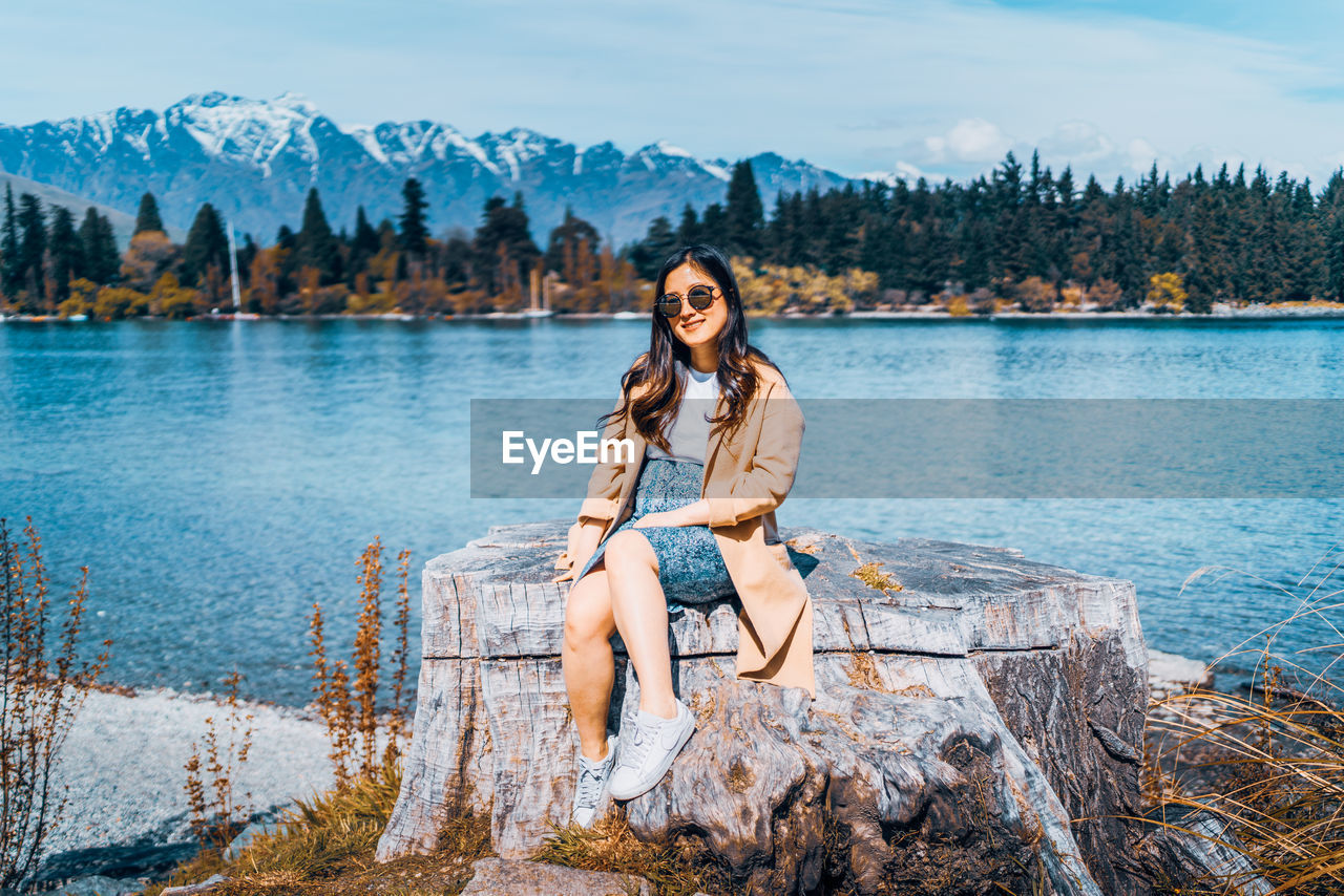 Portrait of young woman sitting on tree stump by lake against mountains and sky