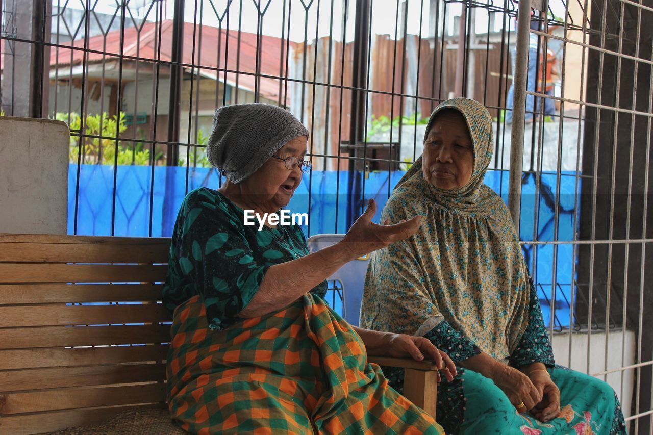 Women talking while sitting on bench against metal grate