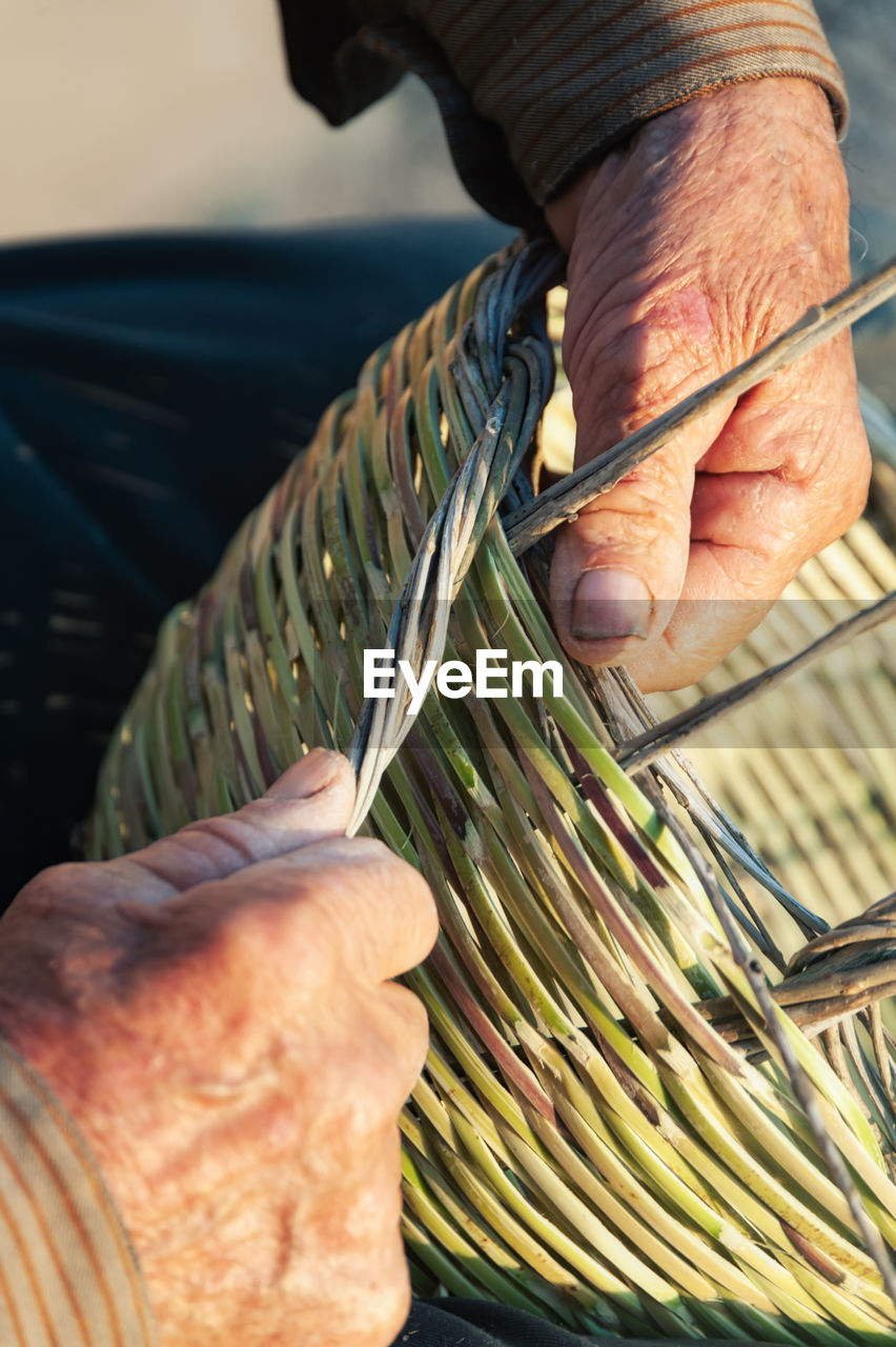 Unrecognizable elderly craftsman making authentic wicker basket on sunny day