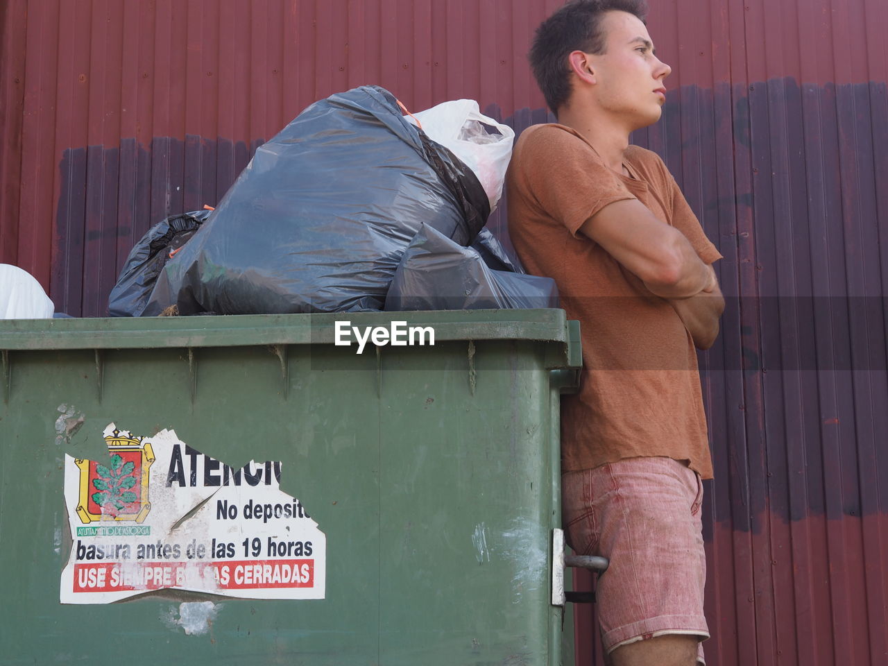 Side view of thoughtful young man standing with arms crossed by garbage bin