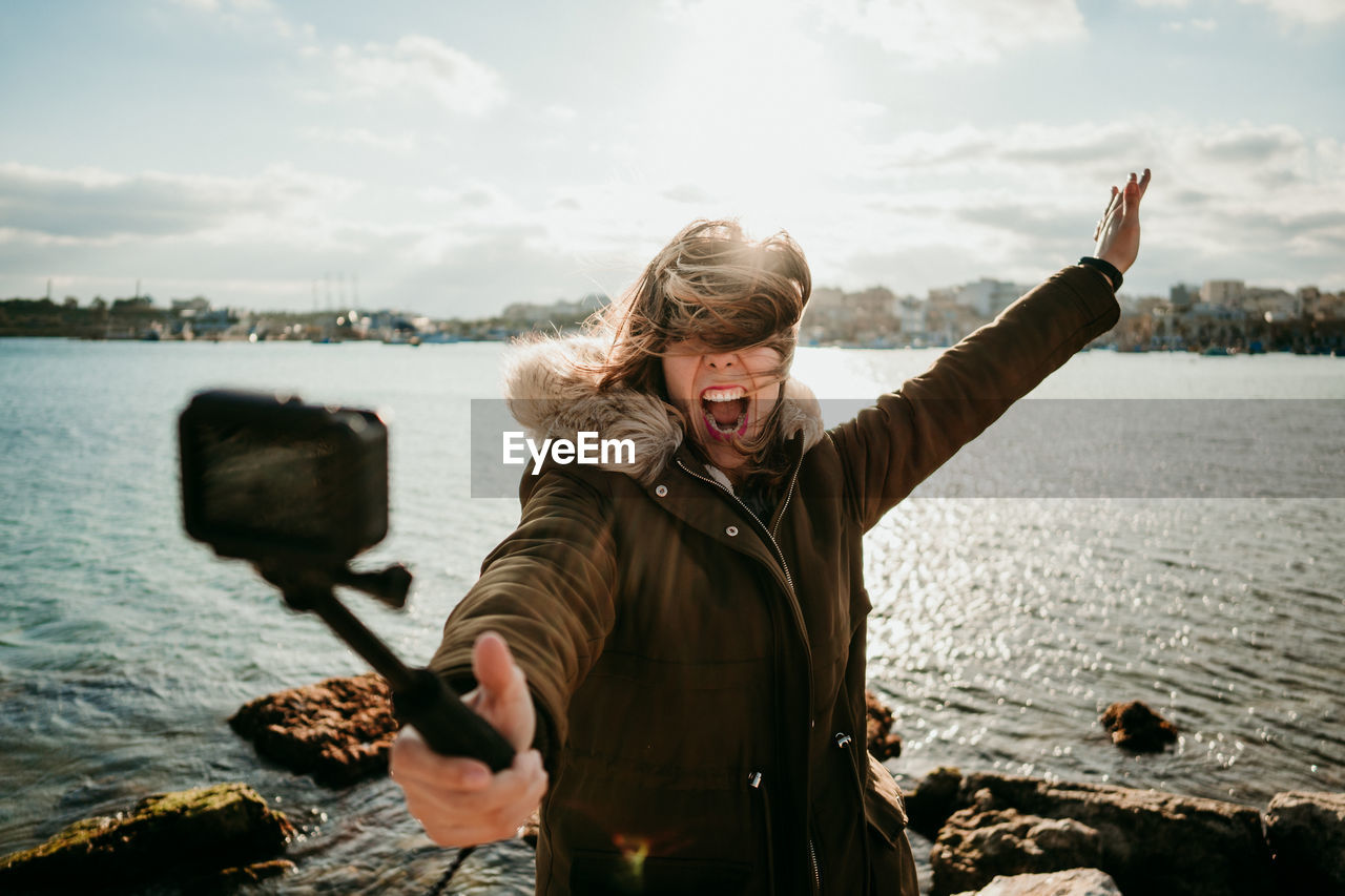 Woman screaming while taking selfie with camera against lake