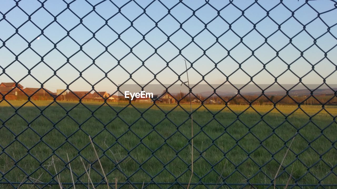 FENCE ON FIELD AGAINST SKY SEEN THROUGH METAL GRATE AGAINST CLEAR BLUE CHAINLINK