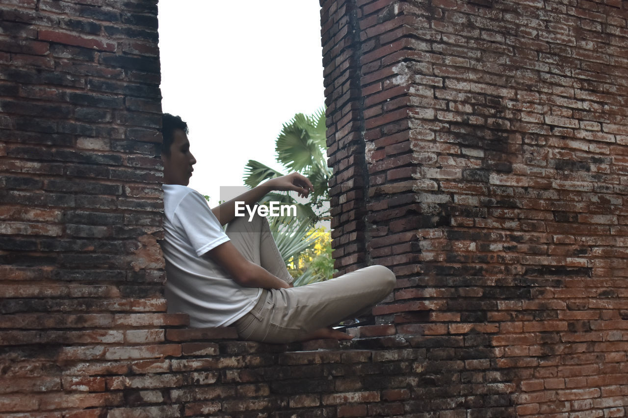 Side view of man sitting on brick wall
