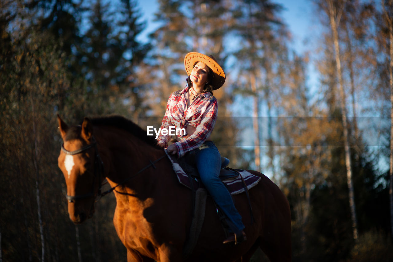 A woman wearing a cowboy hat rides a horse in a countryside farm yard,natural soft sunlight