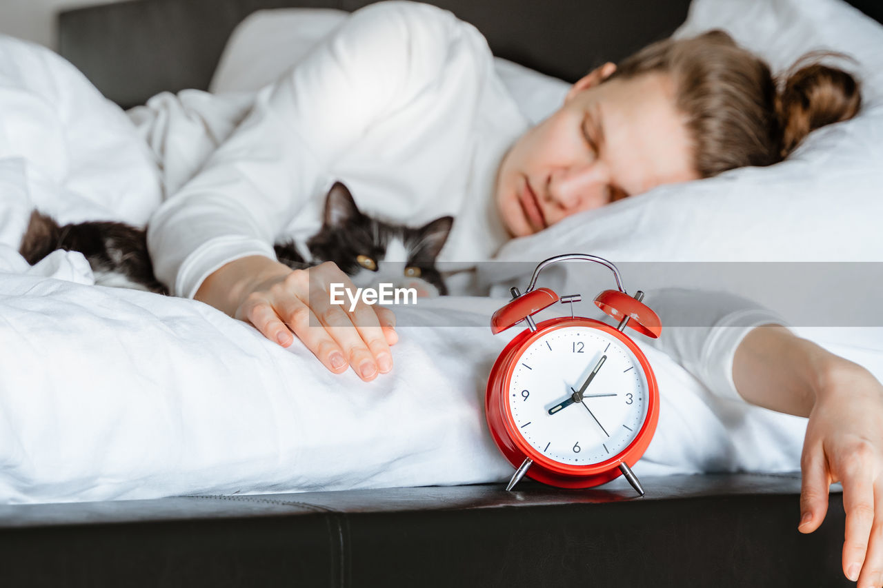 Young millennial woman sleeping with pet cat on white blanket bed cozy at home. alarm clock,