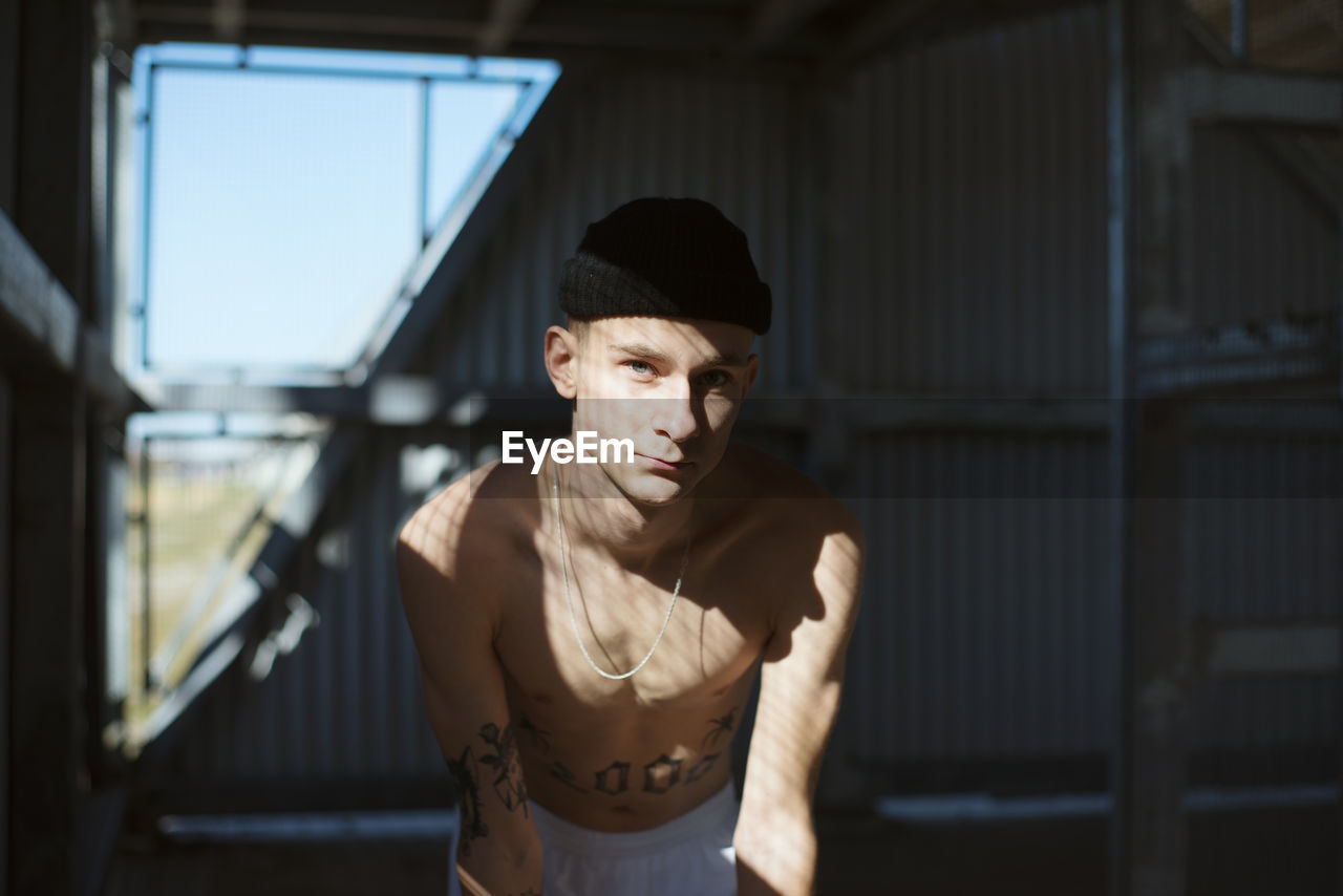 Portrait of shirtless young man wearing knit hat standing indoors