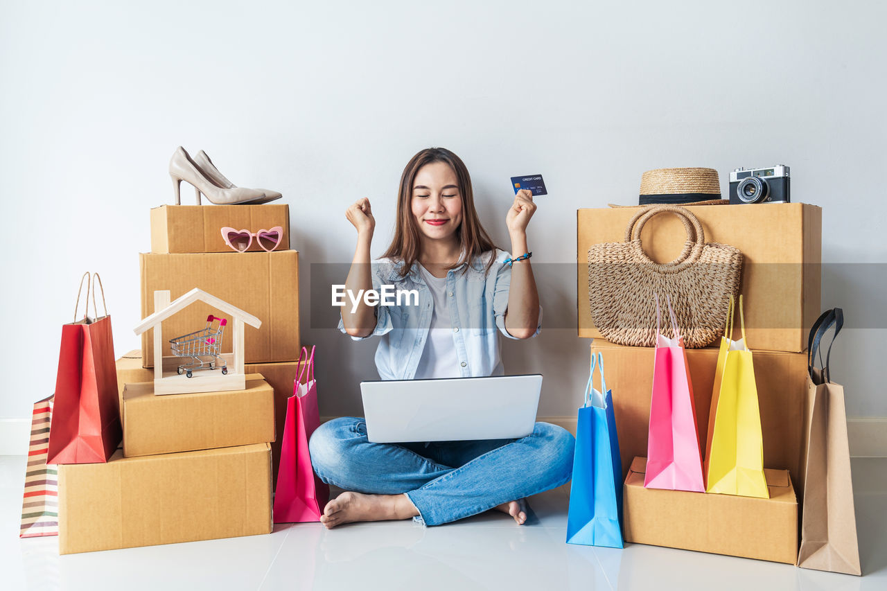 FULL LENGTH OF YOUNG WOMAN USING PHONE WHILE SITTING ON BOX