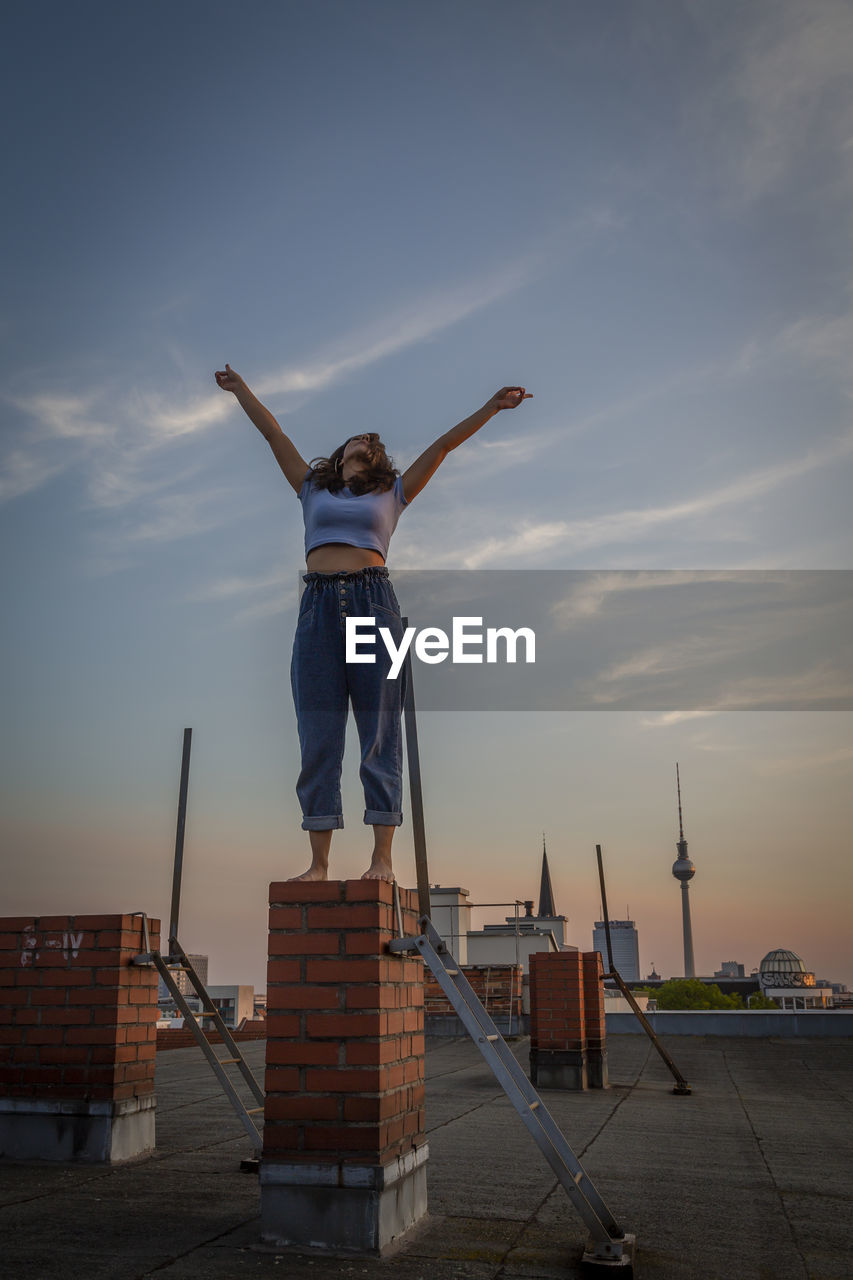 Happy woman with arms raised standing on rooftop against sky during sunset