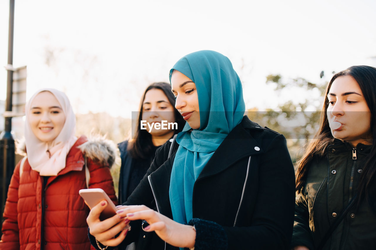 Young muslim woman using smart phone while standing with female friends against sky