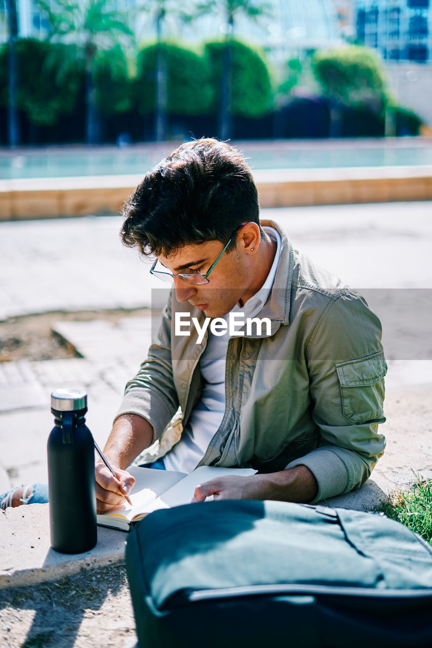 Focused male student sitting on stone bench in university campus and writing in notepad while doing homework on sunny day