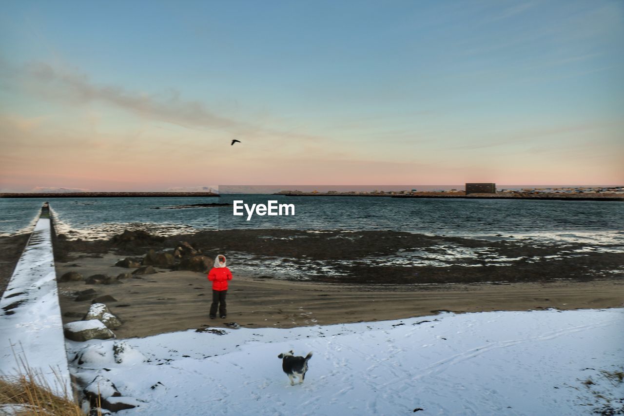 MAN WITH DOG STANDING ON BEACH AGAINST SKY DURING SUNSET