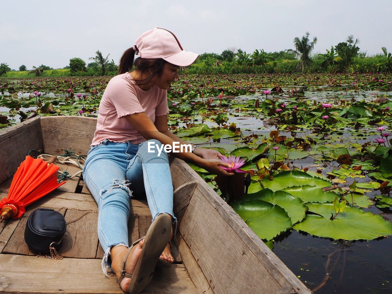 Full length of young woman touching lotus water lily while sitting in boat against sky