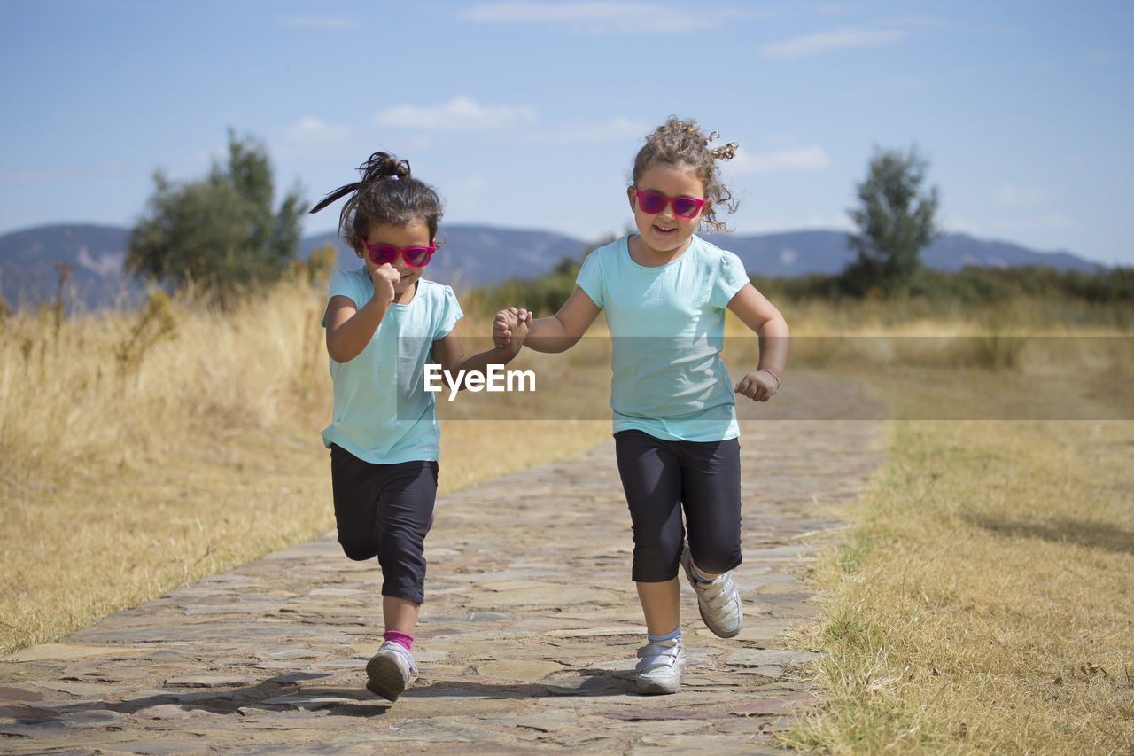 Full length of sisters holding hands while running on footpath against sky