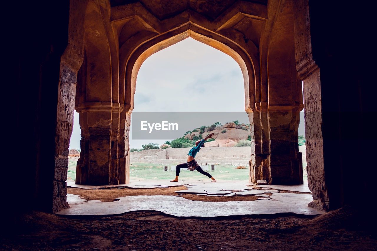 Side view of unrecognizable fit female in activewear performing warrior i pose while practicing yoga near old arched oriental palace in mountainous terrain