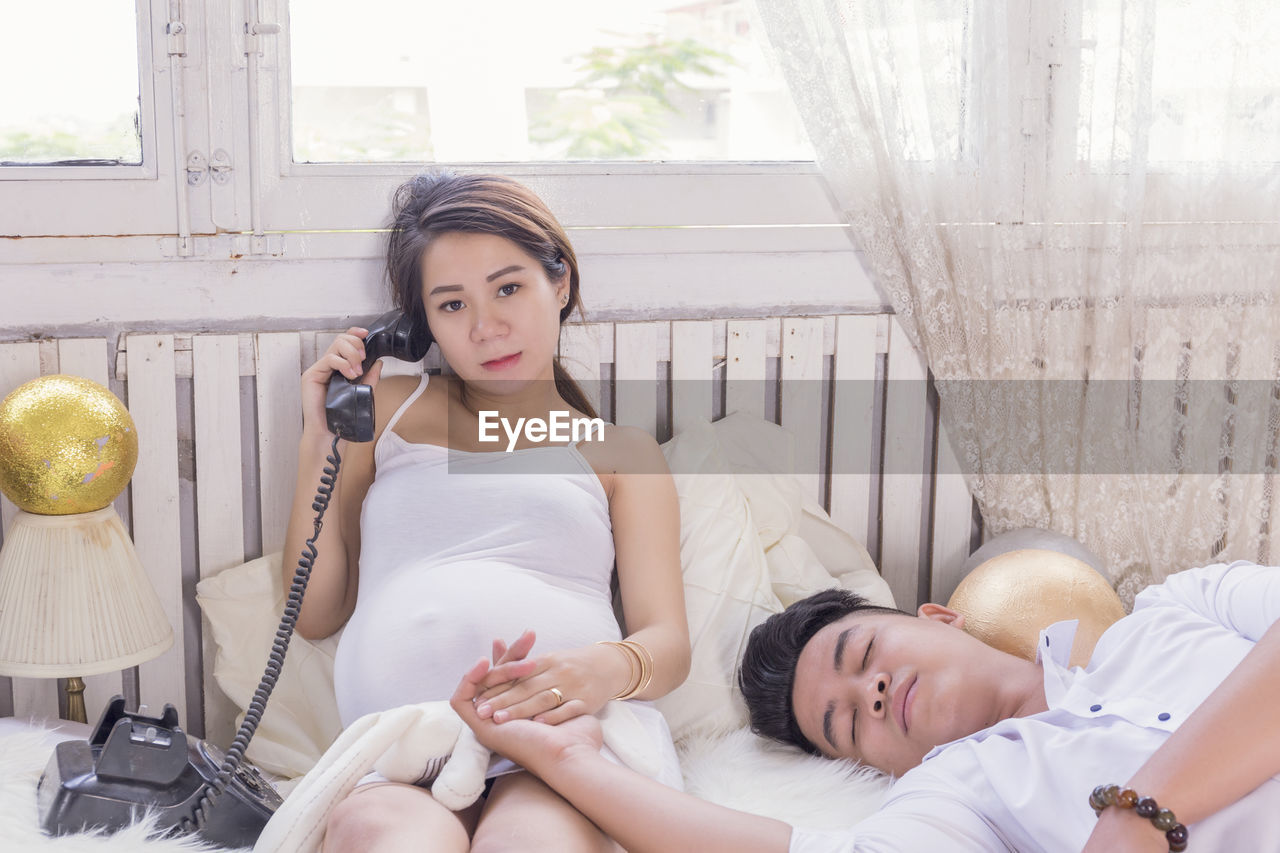 Portrait of pregnant woman talking on phone with boyfriend sleeping on bed at home