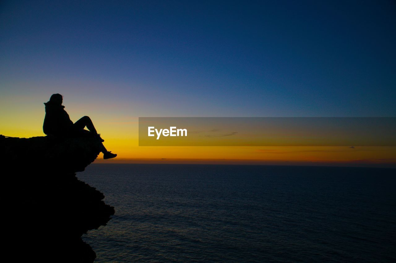 Silhouette woman sitting on rock by sea against sky during sunset