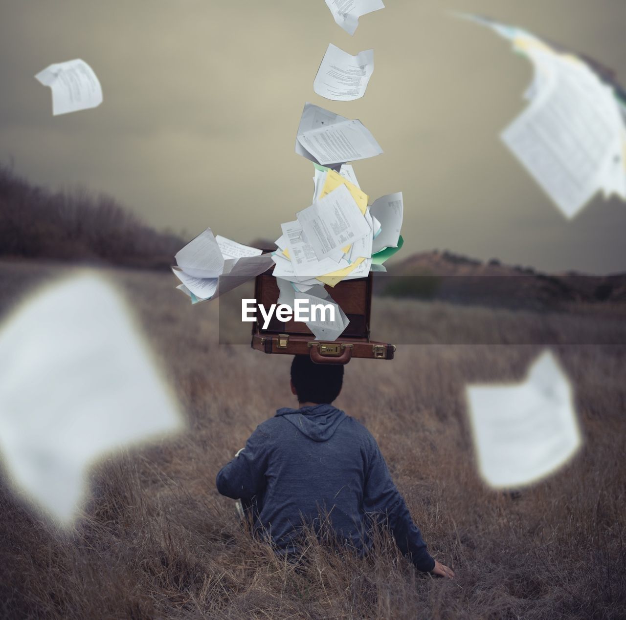 Digitally composite image of papers coming out from briefcase while man sitting on field
