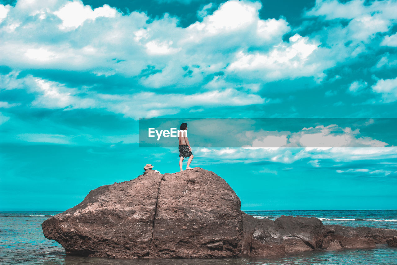 Shirtless man looking at sea while standing on rock against blue sky