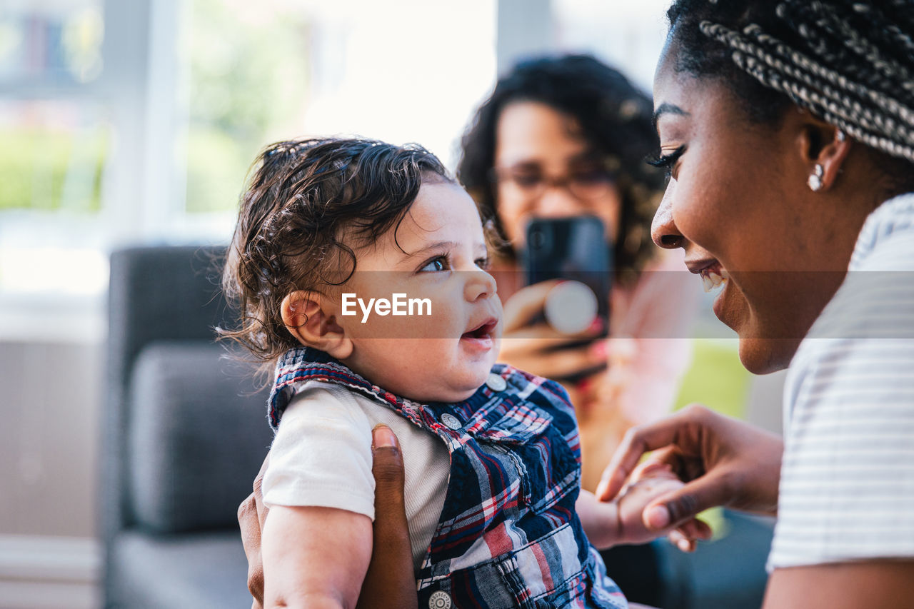 Cheerful mother playing with cute son while girlfriend photographing them in living room