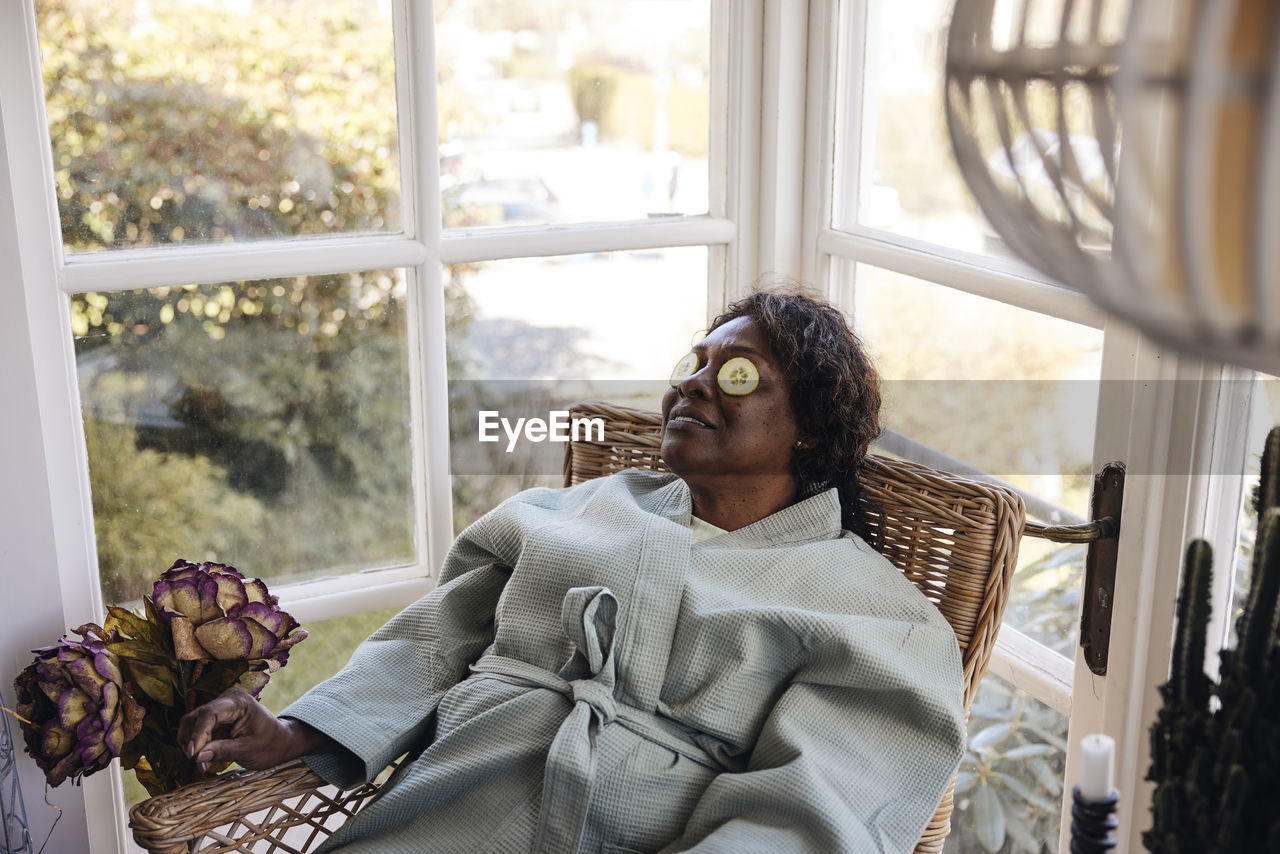 Mature woman with cucumber slices on eyes sitting on chair at home