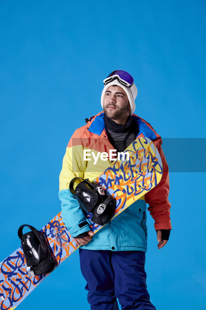 adult, sports, one person, blue, extreme sports, men, snowboarding, skiing, clothing, sky, snowboard, three quarter length, young adult, leisure activity, emotion, standing, portrait, nature, low angle view, mountain, winter sports, winter, day, clear sky, athlete, sports equipment, copy space, colored background, sports clothing, ski goggles, looking at camera, outdoors