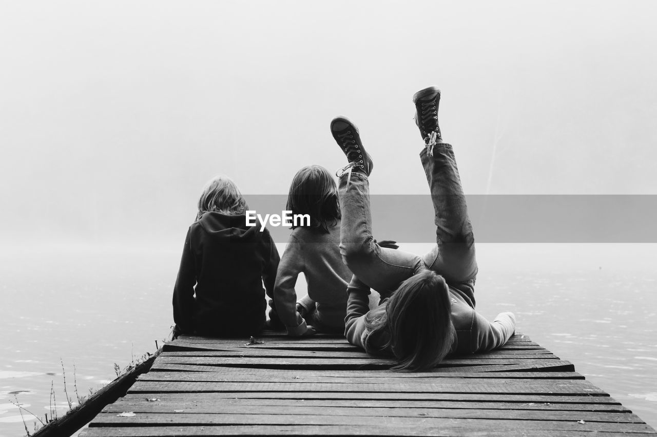 Rear view of friends with arm around sitting on pier by river during foggy weather