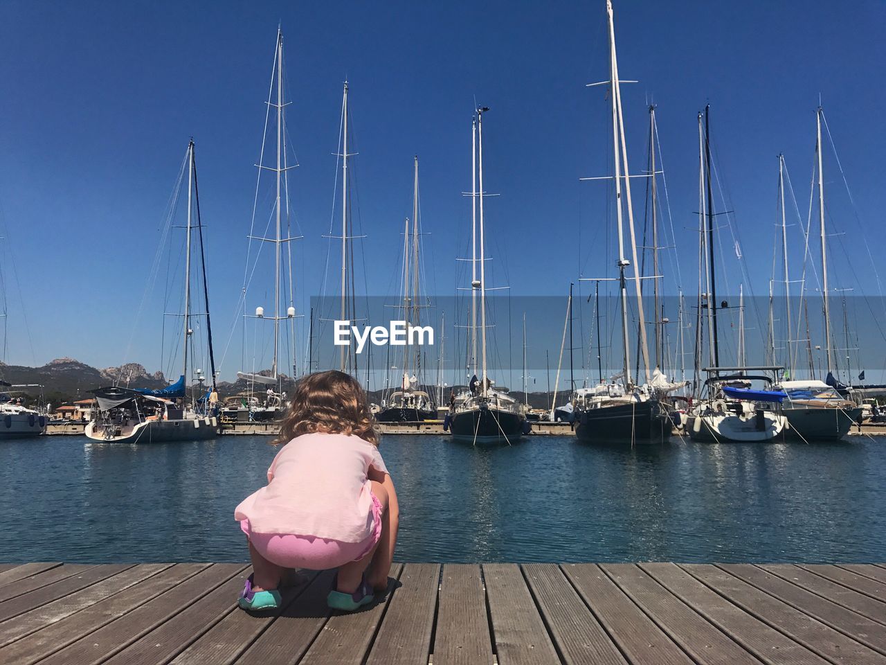 Rear view of baby girl crouching on pier at harbor with boats in background against clear sky