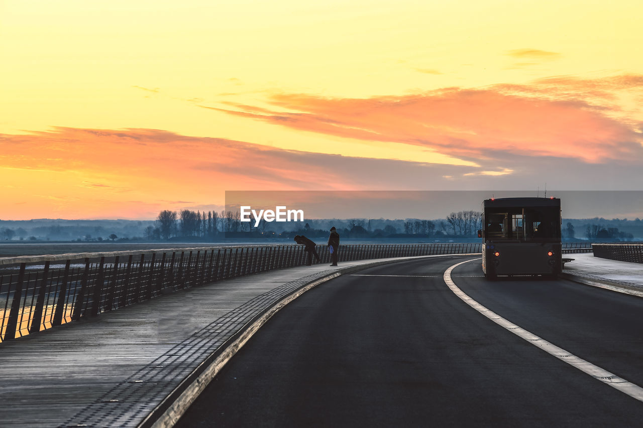 People on sidewalk with vehicle on bridge against sky during sunset