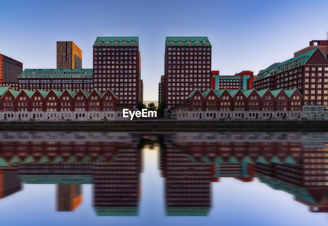 Buildings reflecting on river in city against sky during sunset