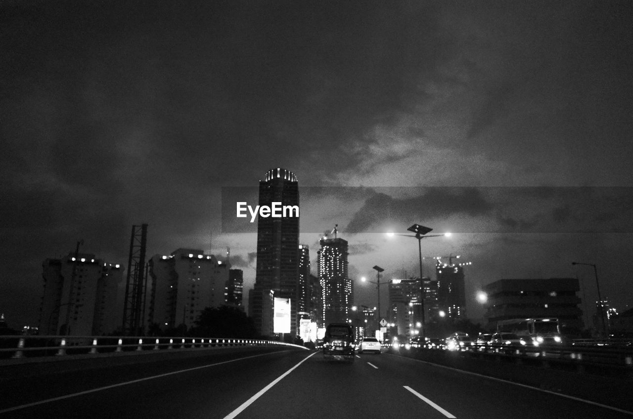 Road leading towards illuminated cityscape against cloudy sky at night