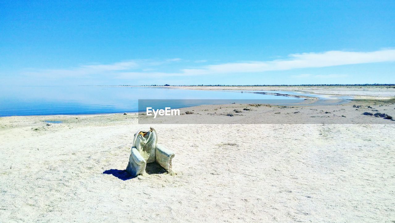 A battered and weather worn armchair sits lonely on beach of salton sea against blue sky