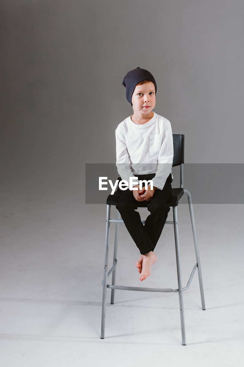 A boy in a white t-shirt and cap sits on a bar stool, on a white background. 