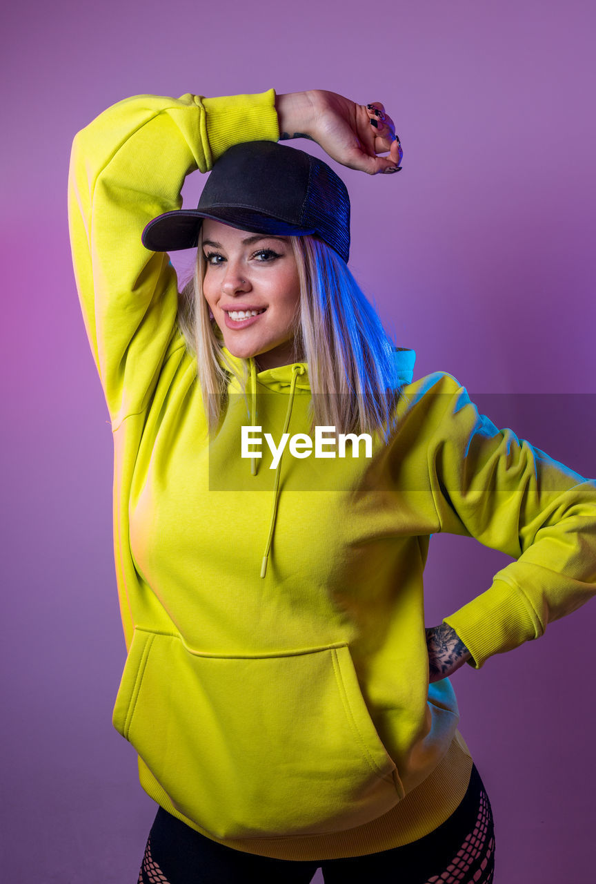 Female in street style hoodie and cap looking at camera on purple background in studio with neon illumination