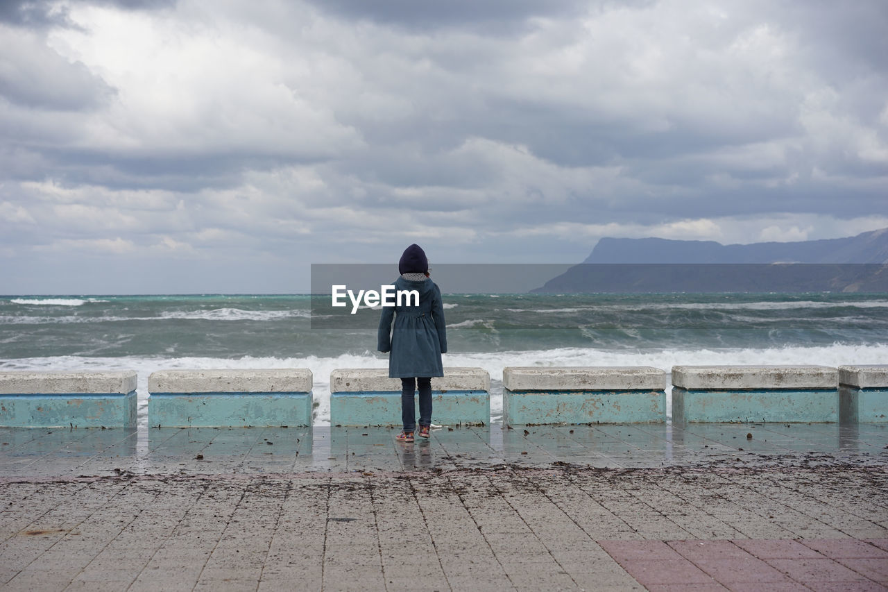 Rear view of woman standing on promenade by sea against cloudy sky