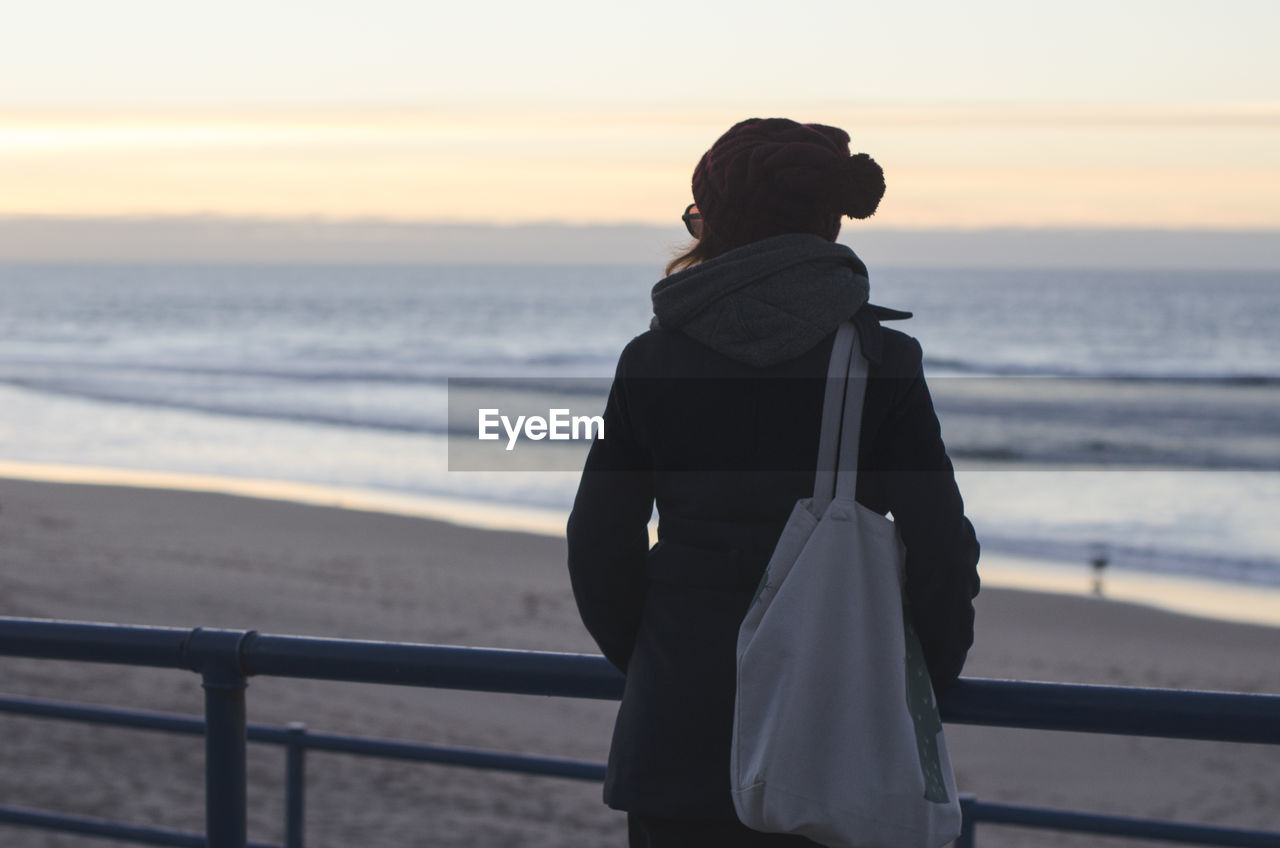 Rear view of woman standing by railing while looking at beach during sunset