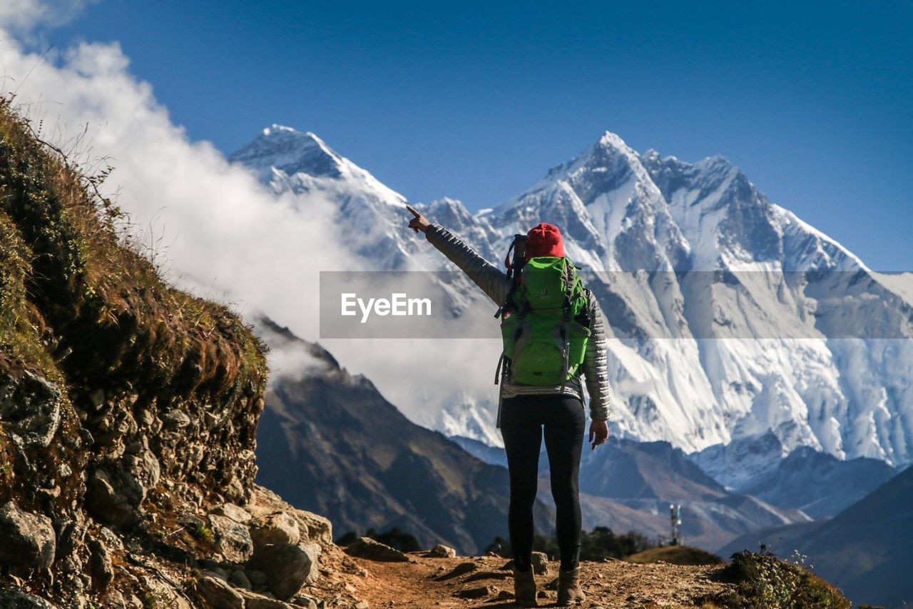 REAR VIEW OF MAN STANDING ON SNOWCAPPED MOUNTAINS AGAINST SKY