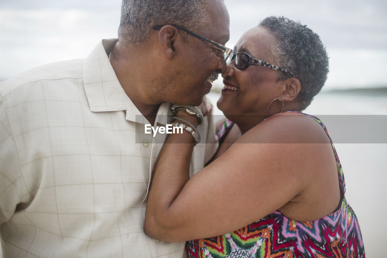 Happy senior couple romancing at beach against cloudy sky