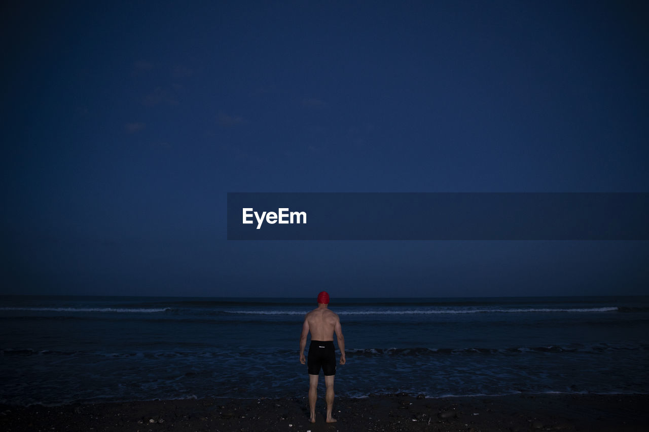 Man in swimming shorts standing at water's edge during dawn