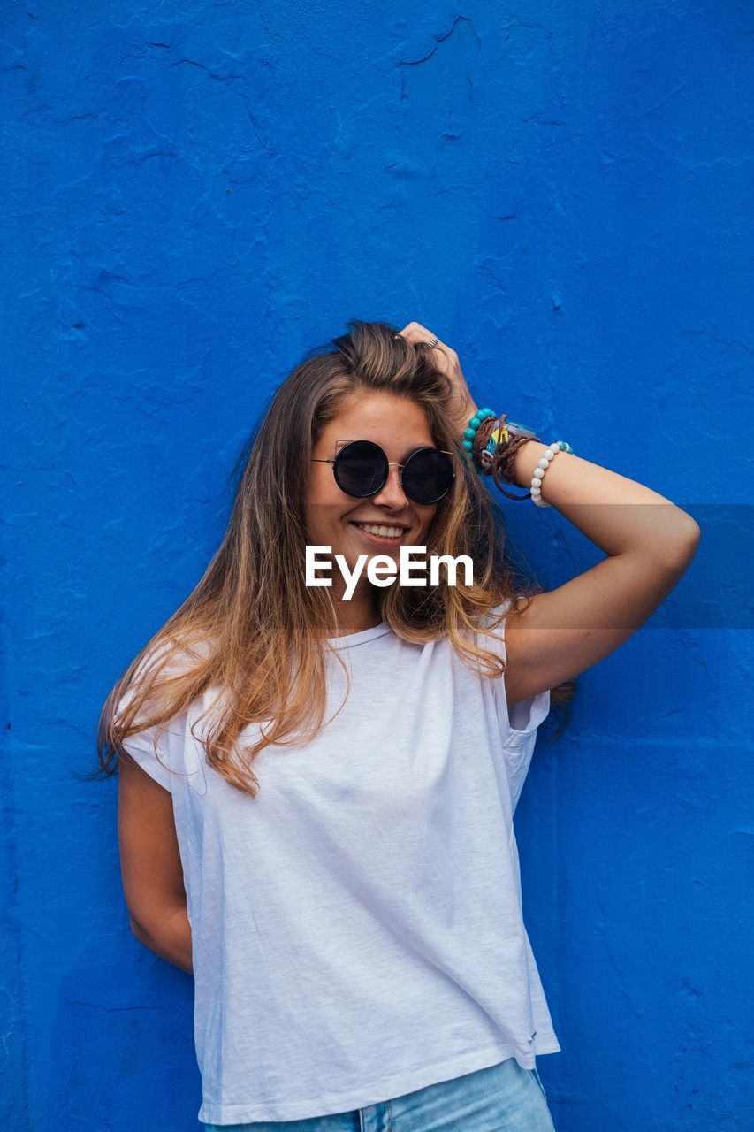 Portrait of smiling young woman in sunglasses standing against blue wall