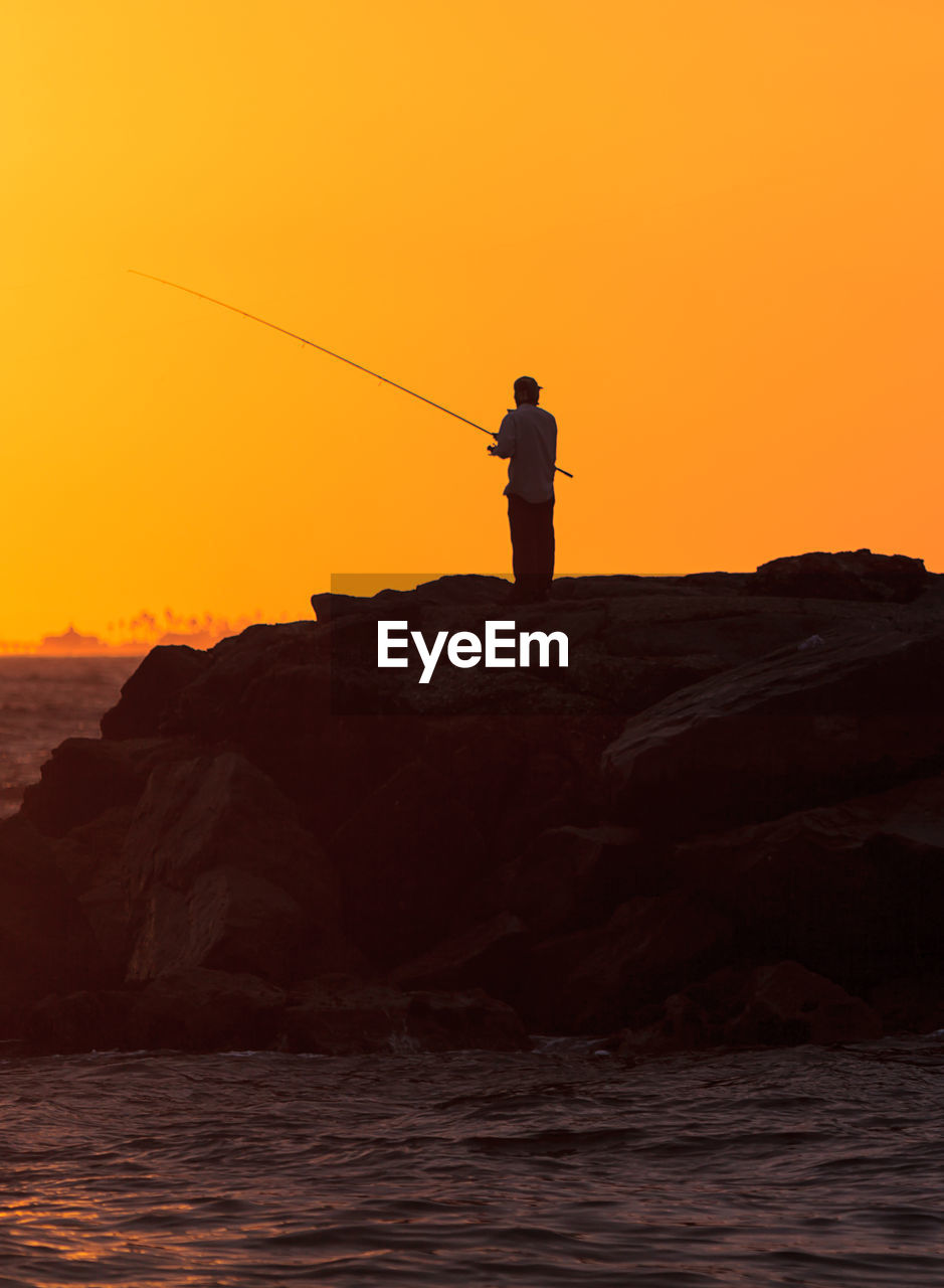 Man fishing while standing on rock by sea against orange sky