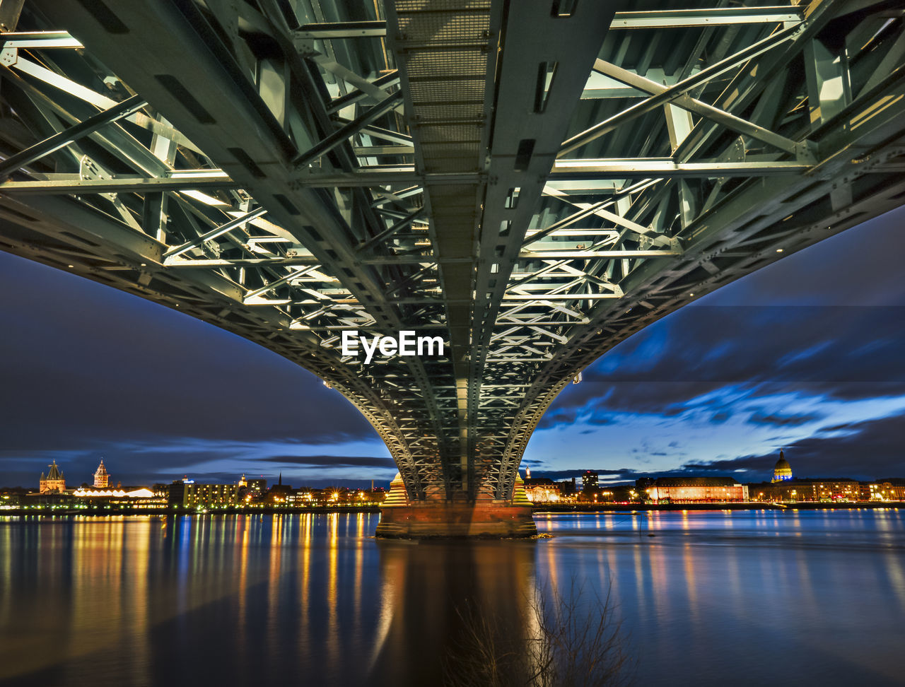 Underneath view of illuminated bridge over river in city at dusk